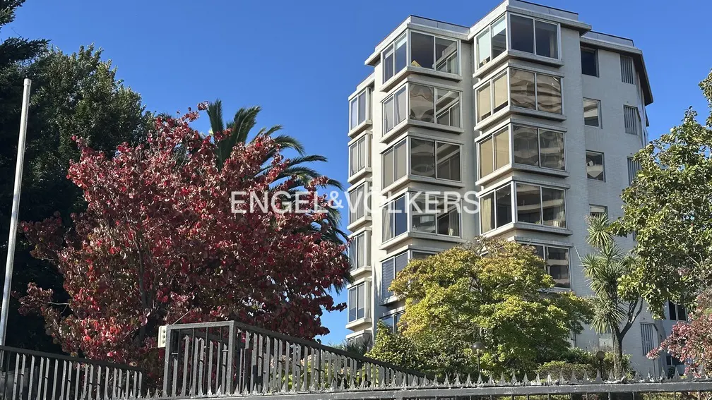 A modern, multi-story apartment building with large windows, surrounded by trees and a black iron fence.