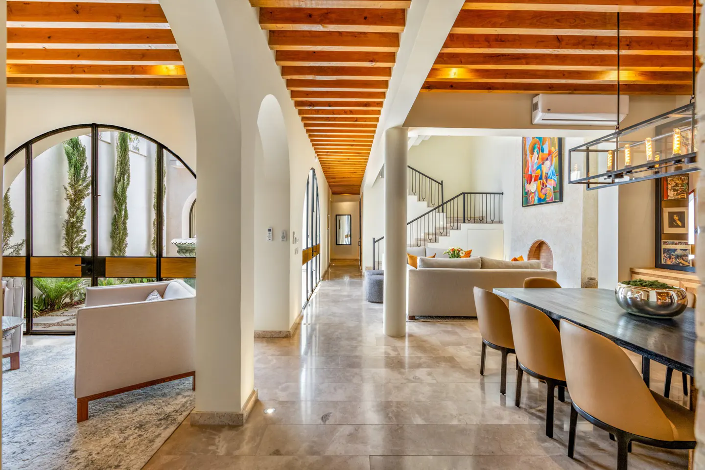 Interior view of a bright, modern home with arched doorways, wood beam ceilings, and marble floors. A dining table with tan chairs is in the foreground.