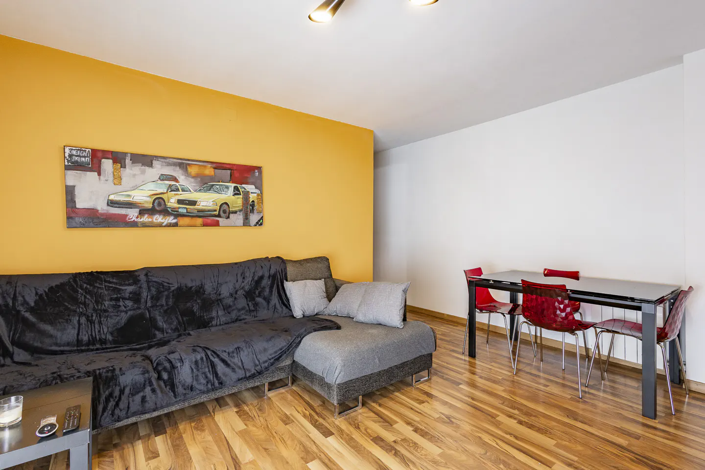 Living room with a black sofa, yellow wall art, and a dining table with red chairs on a wood floor.