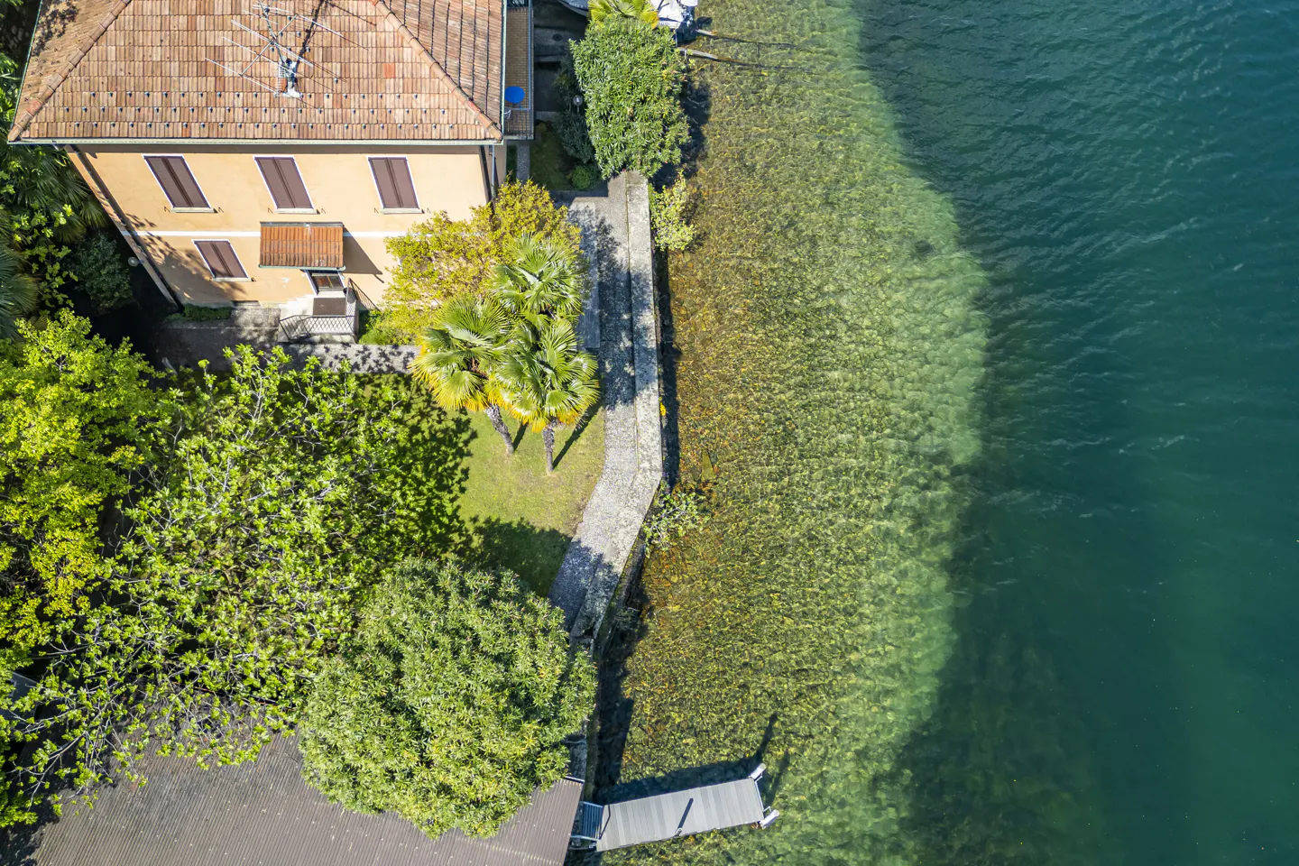 Aerial view of a tan house with a red tile roof next to a clear lake with a dock.