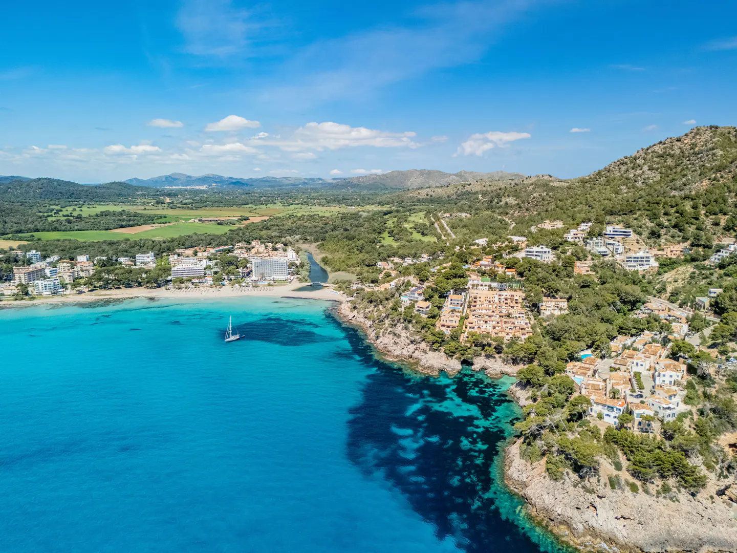 Aerial view of a turquoise bay with a sailboat, sandy beach, and hillside villas under a blue sky.