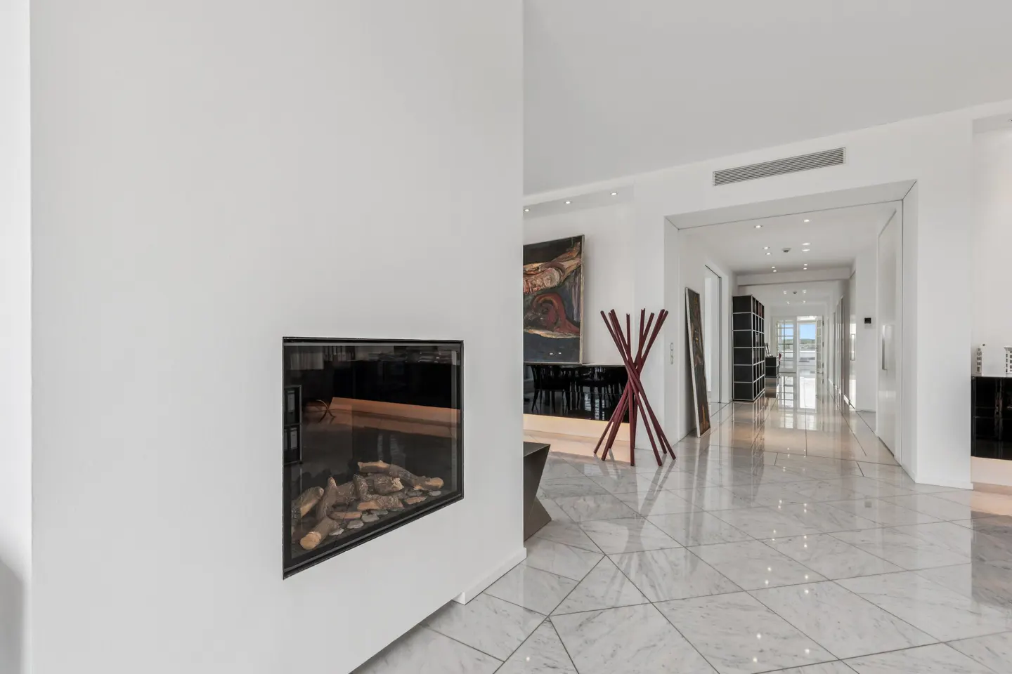 Bright, modern hallway with marble floors. A fireplace is set into a white wall. A red coat rack stands near an abstract painting.