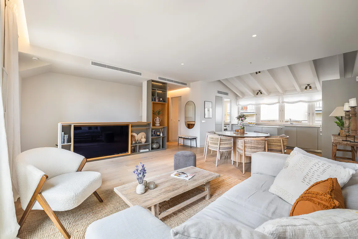 Modern open-plan living room with a white sofa, cream armchair, wooden coffee table, and dining area with wicker chairs.