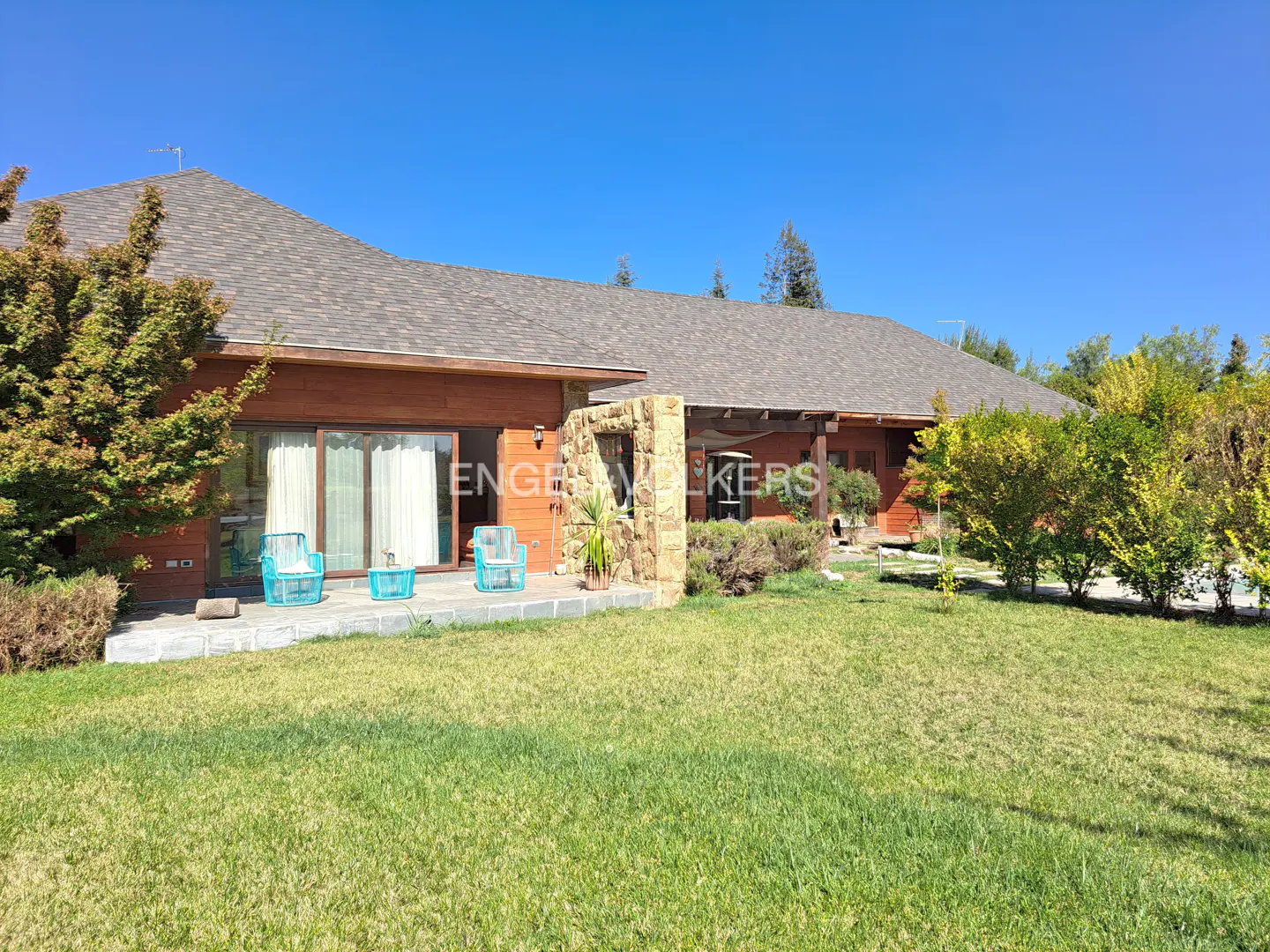 Exterior view of a wood-sided house with a stone accent wall and a large lawn under a clear blue sky. Patio furniture is visible.