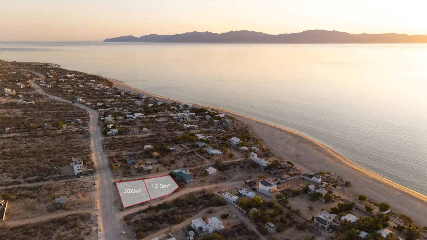 Aerial view of two adjacent lots, outlined in red, sized 1,026m² and 1,018m², near a beach with mountains in the distance.