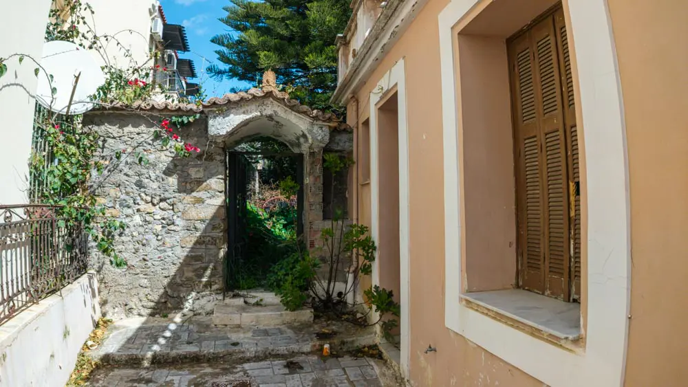 Stone archway leads to a garden path. Peach-colored building with brown shutters on the right. Vines and flowers on the left.