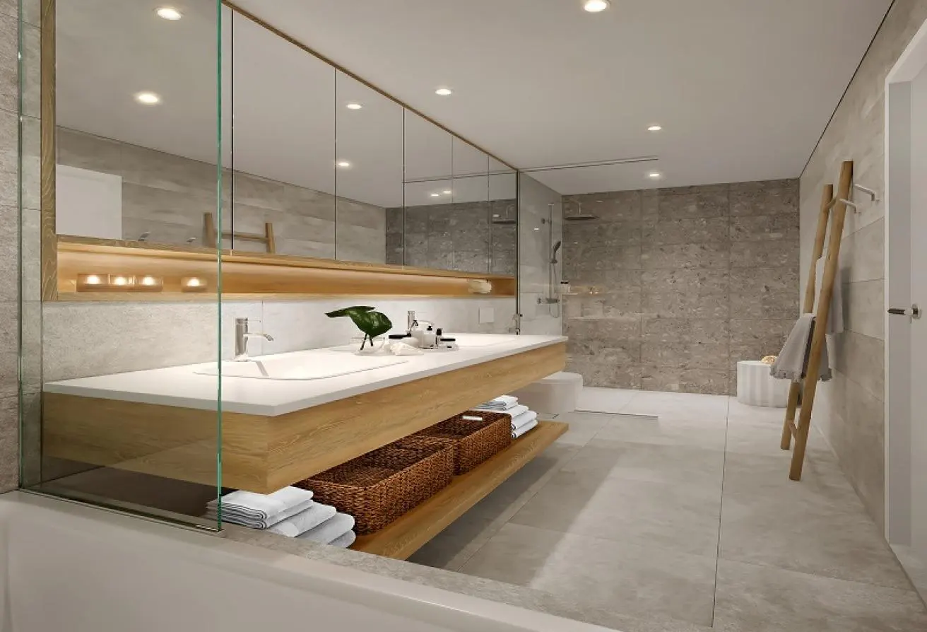 A modern bathroom with a long, light wood vanity, white countertop, and large mirror. Gray tile walls, a wooden towel ladder, and recessed lighting complete the space.