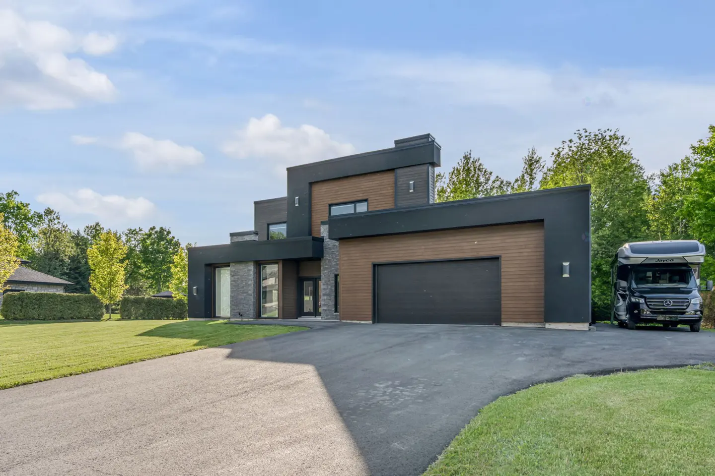 Modern two-story home with black and wood siding, stone accents, a large garage, and an RV parked on the asphalt driveway.