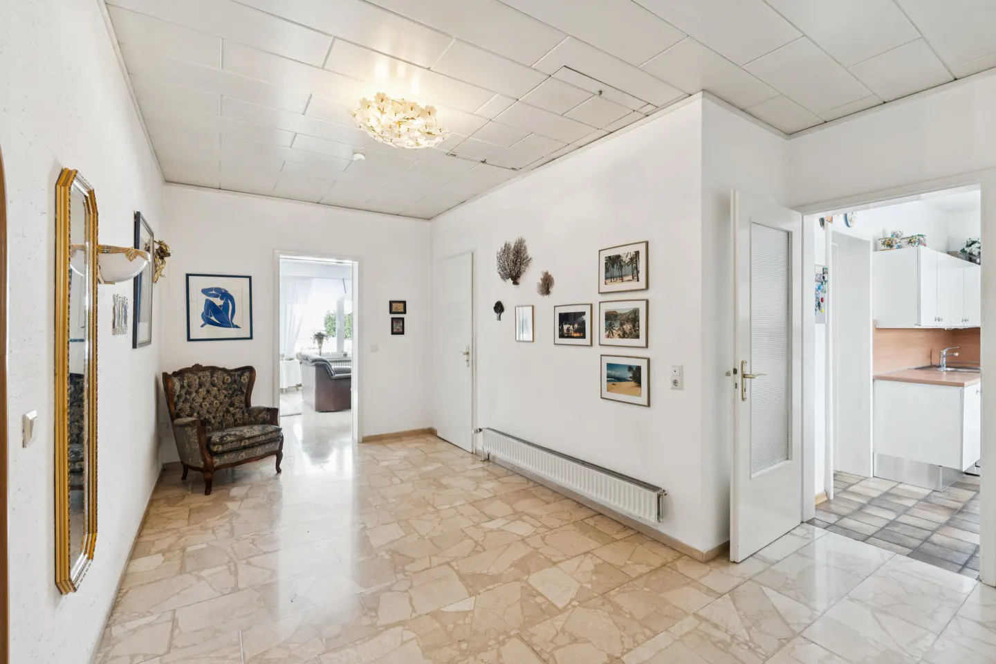 A bright hallway with marble floors, white walls, and a decorative mirror. A patterned armchair sits near a doorway, with framed art on the walls.