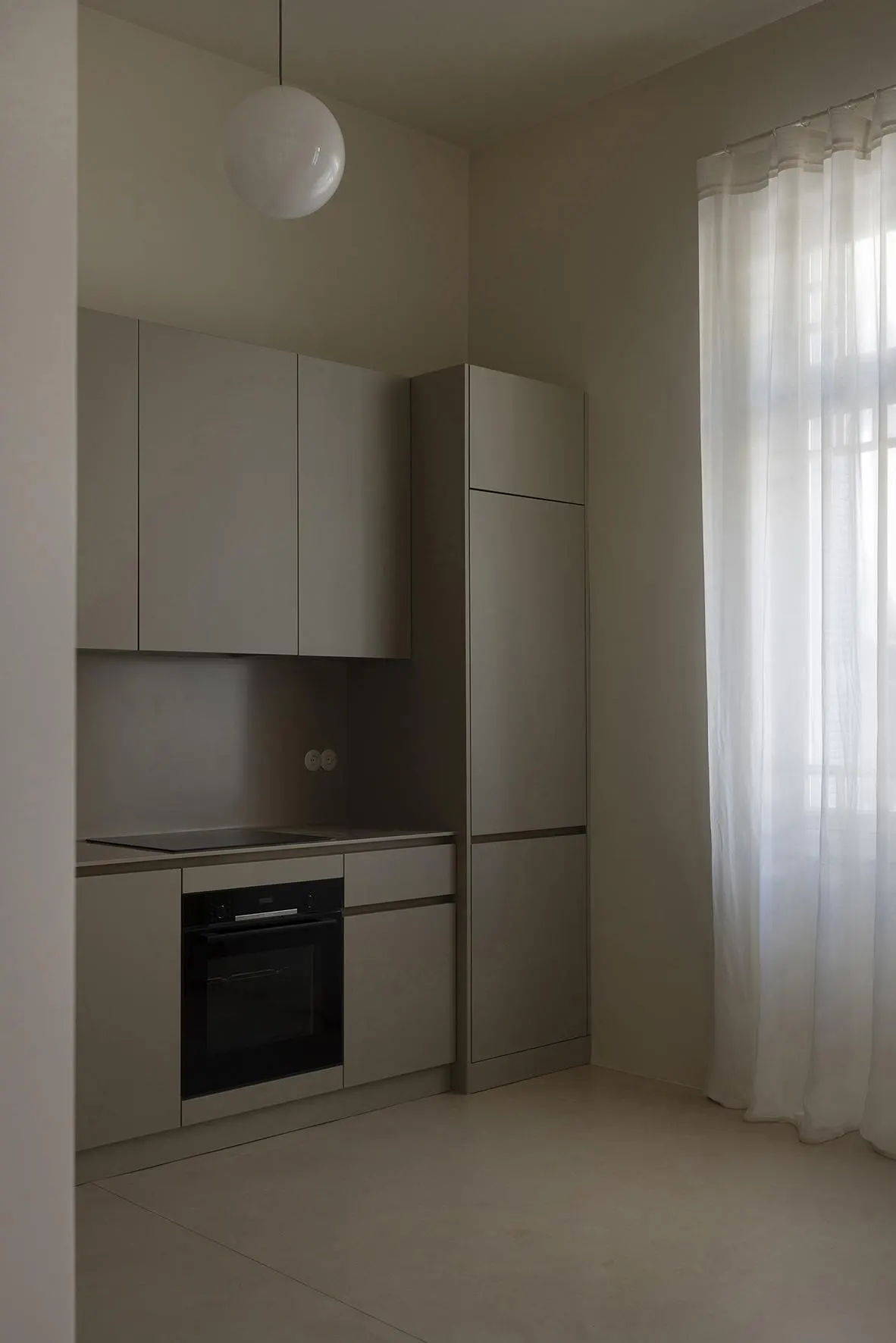 A modern kitchen with gray cabinets, black oven, and white pendant light. A window with sheer white curtains is on the right.