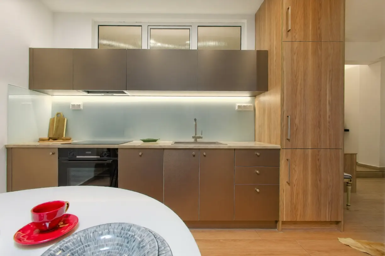 Modern kitchen with matte brown cabinets, wood-grain pantry, light backsplash, and a white table with a red cup in front.