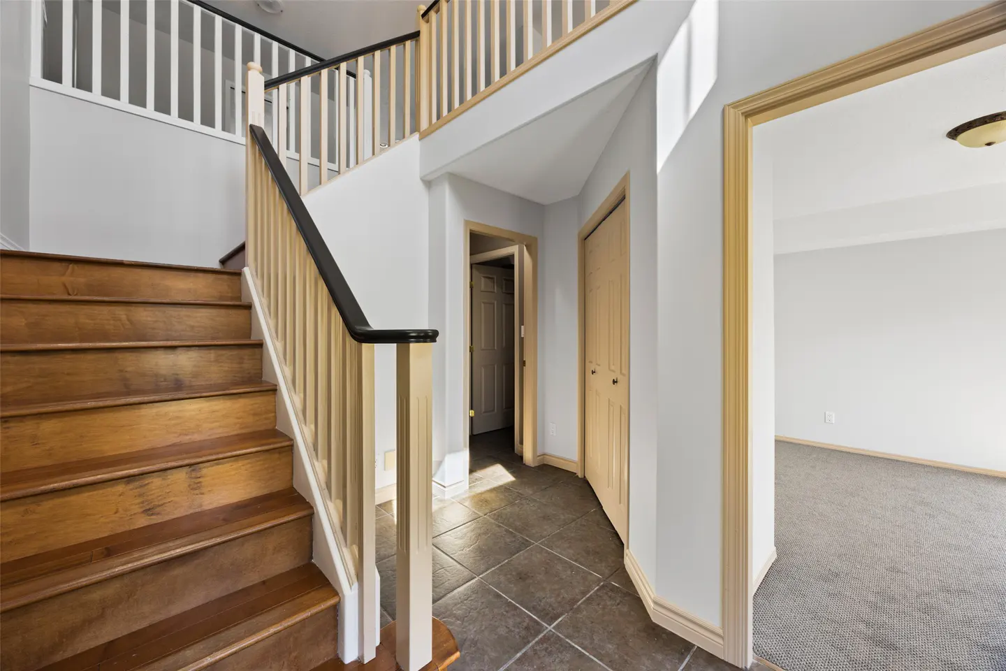 Interior view of a home's foyer with wooden stairs, tile floor, and doorways leading to other rooms. Walls are light gray.