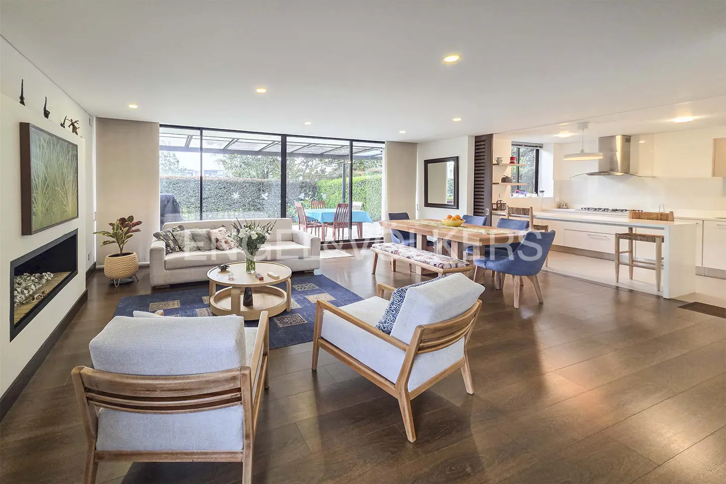 Open-concept living space with hardwood floors, a white sofa, chairs, and a dining table. Large windows offer a view of the outdoor patio.