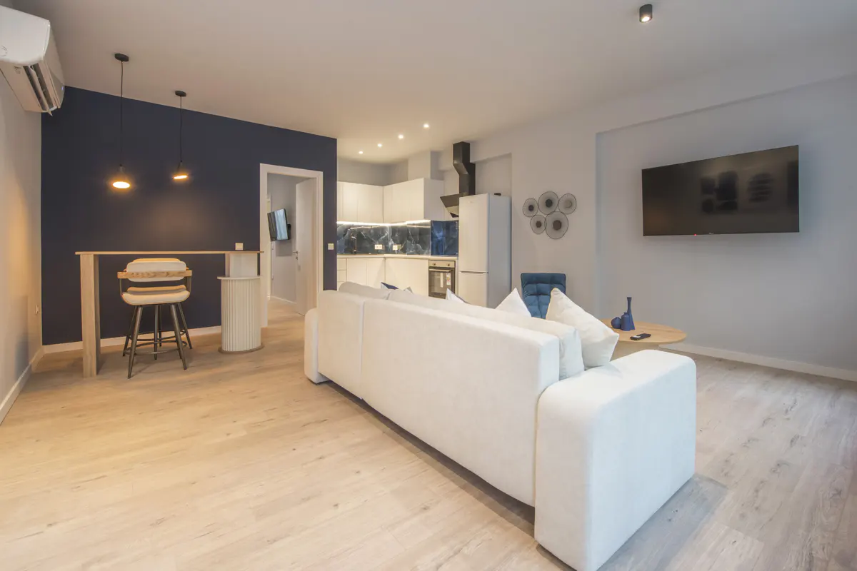 Bright, modern apartment interior with a white sofa, bar stools, and a kitchen in the background. A large TV is mounted on the wall.