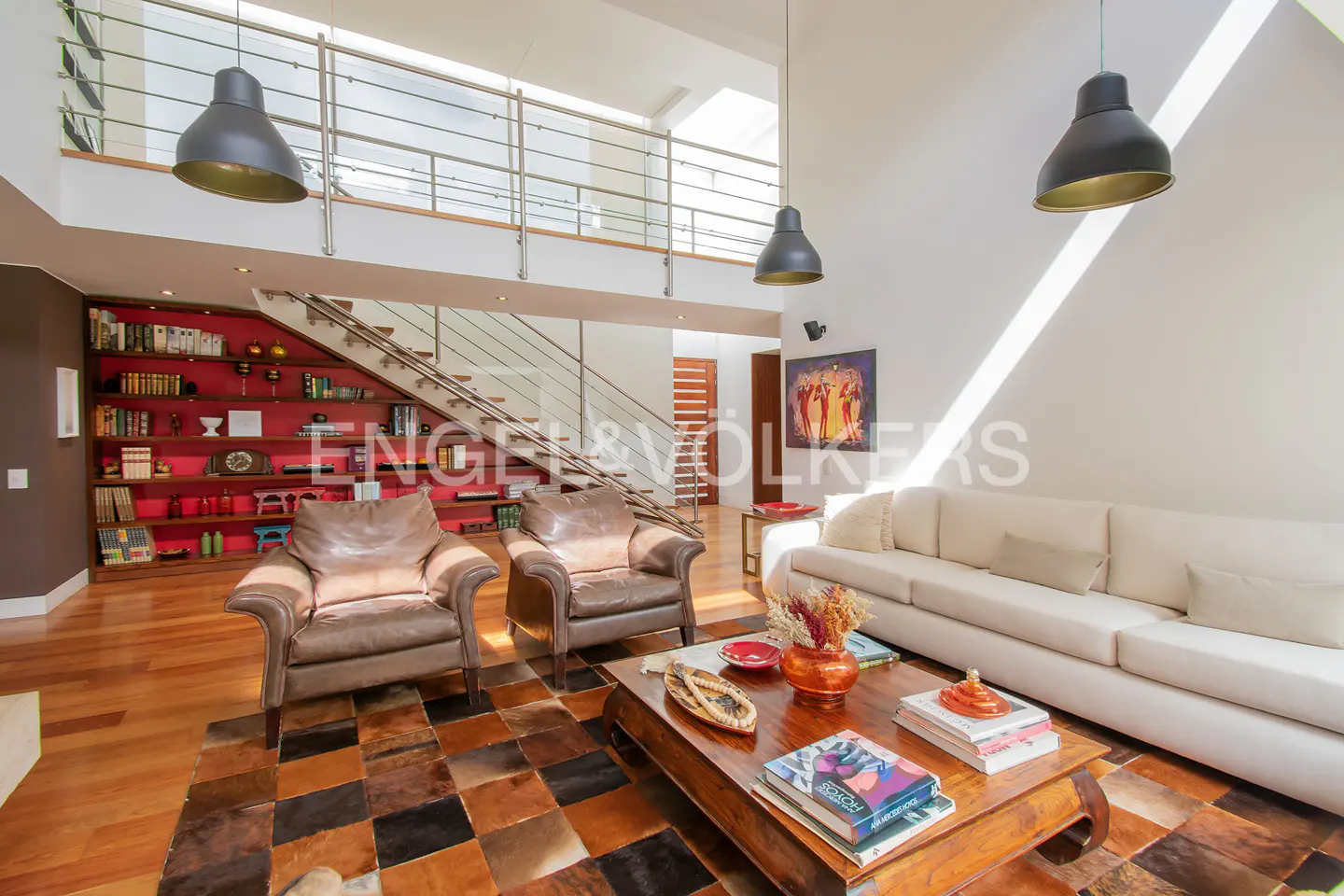 Modern living room with high ceilings, leather armchairs, white sofa, wooden coffee table, and a red bookshelf staircase.