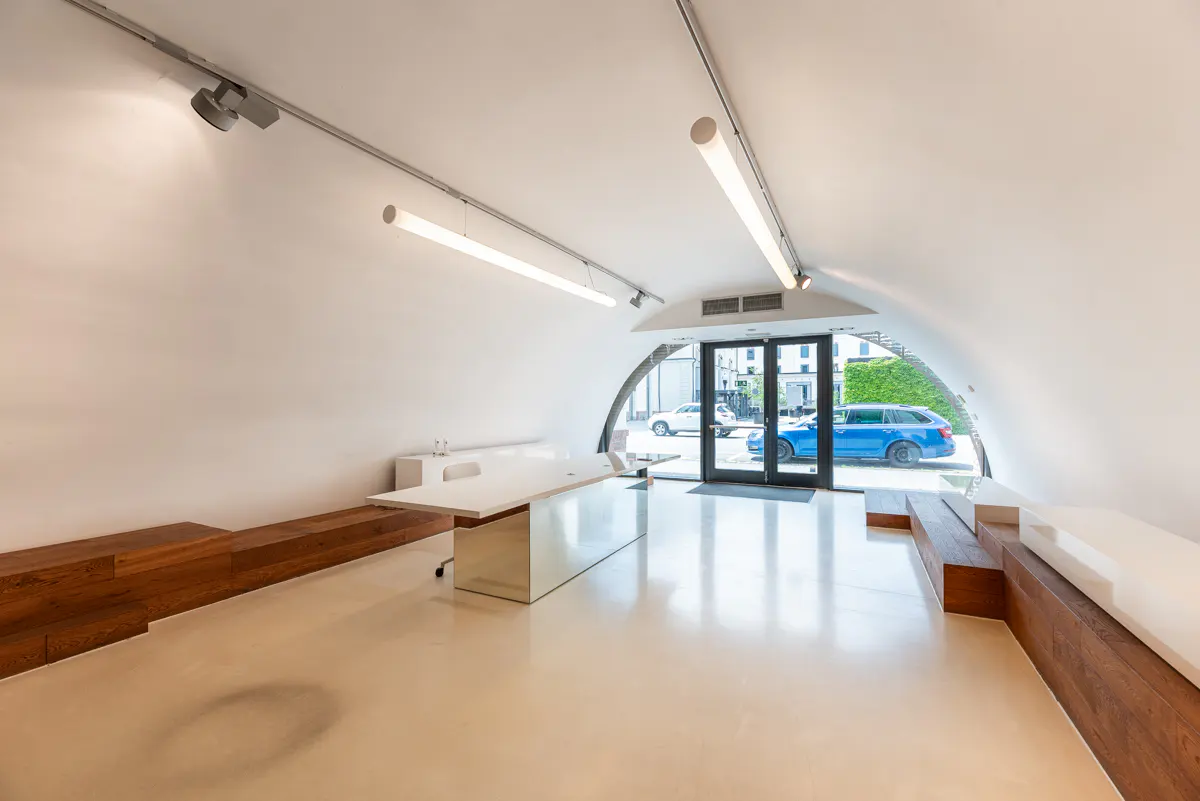 Bright, modern office space with white walls, a curved ceiling, and a large white desk. Wood accents and a glass door leading outside are visible.