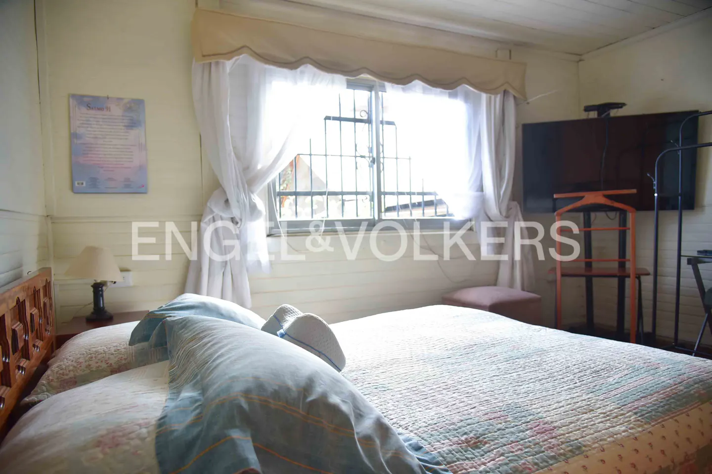 Bedroom with a bed, blue pillows, white curtains, and a window with bars. A TV and a wooden chair are in the background.