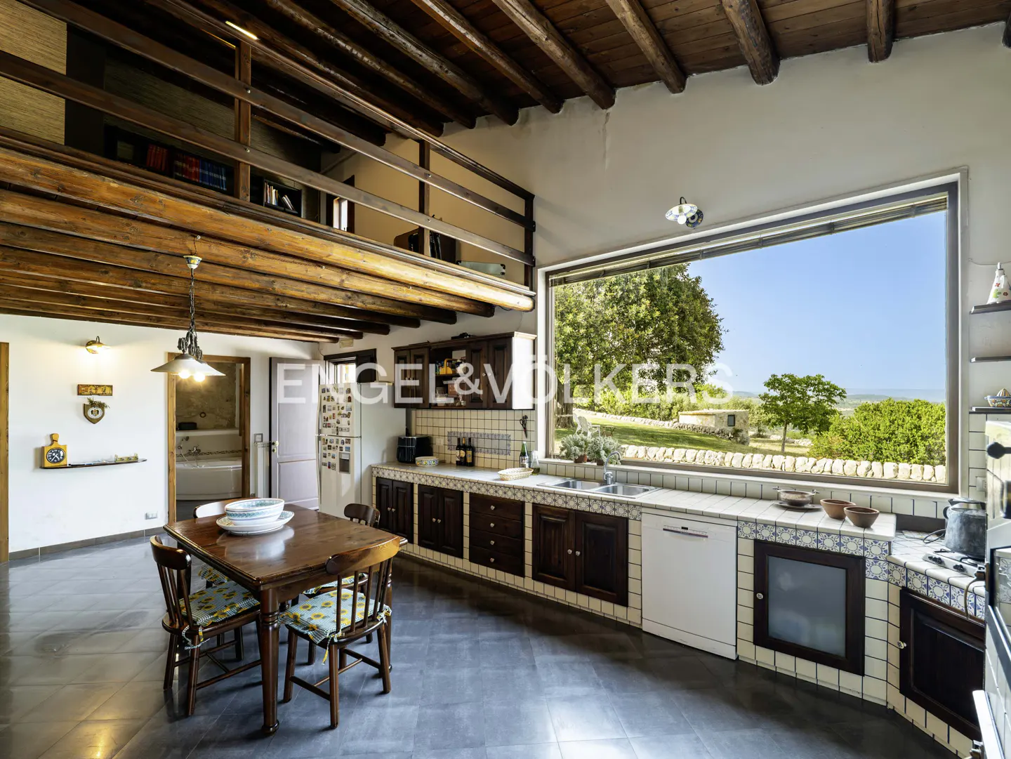 Rustic kitchen with wooden beams, a loft, a dining table, and a large window overlooking a sunny, green landscape.