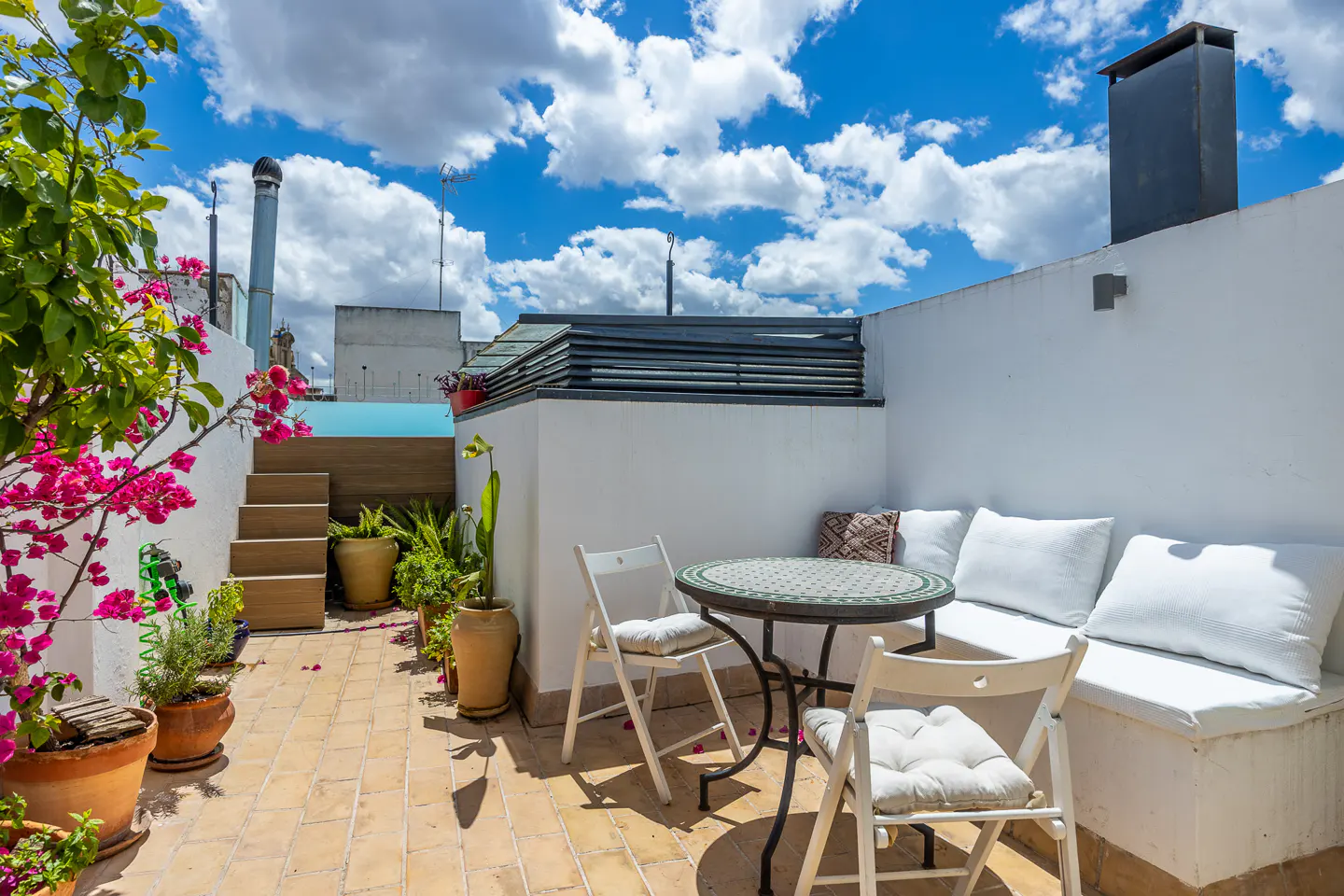 Outdoor patio with white walls, a table and chairs, and potted plants under a blue sky with white clouds.