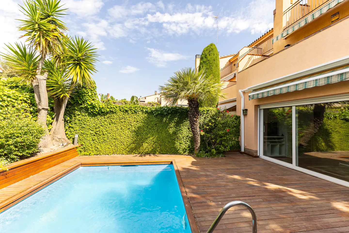 A backyard with a blue pool, wooden deck, and green ivy wall. A tan building with a striped awning is in the background.