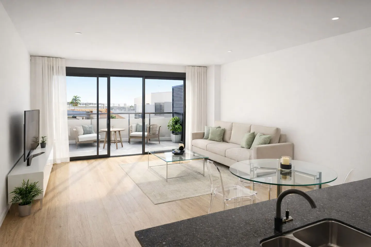 Bright living room with a beige sofa, glass tables, and a balcony with outdoor seating. White walls and light wood floors create a modern, airy feel.