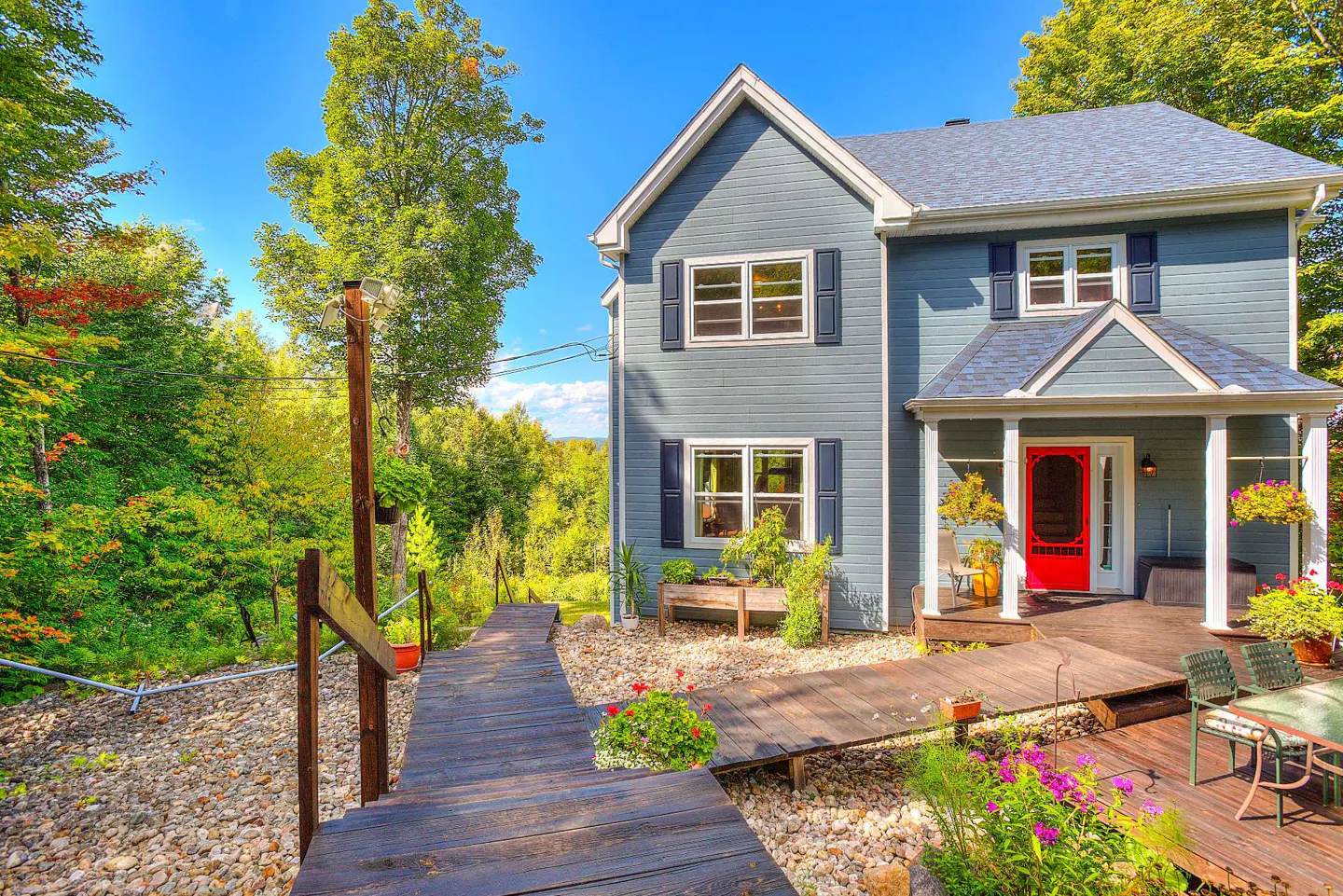 Two-story blue house with a red door, dark blue shutters, and a wooden walkway leading to the front porch. Lush green trees surround the house.