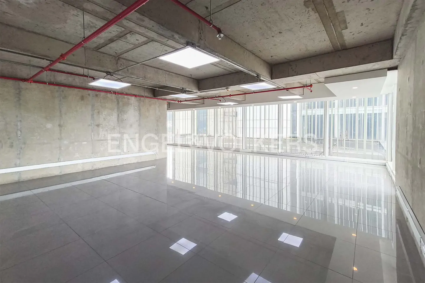 Unfurnished office space with concrete walls, gray tile floor, and large windows. Red pipes run across the ceiling with square LED lights.