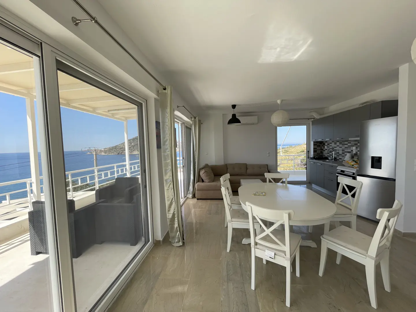 Bright, open-concept living space with a white dining table and chairs. Sliding glass doors lead to a balcony with ocean views. Gray kitchen cabinets are visible.
