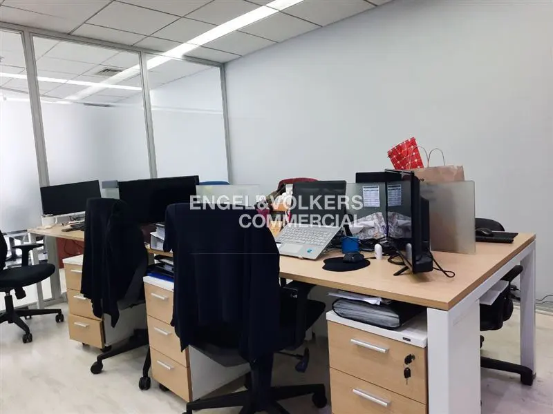 Office space with desks, computers, and chairs. The walls are white, and there is a glass partition. Engel & Volkers Commercial logo is visible.