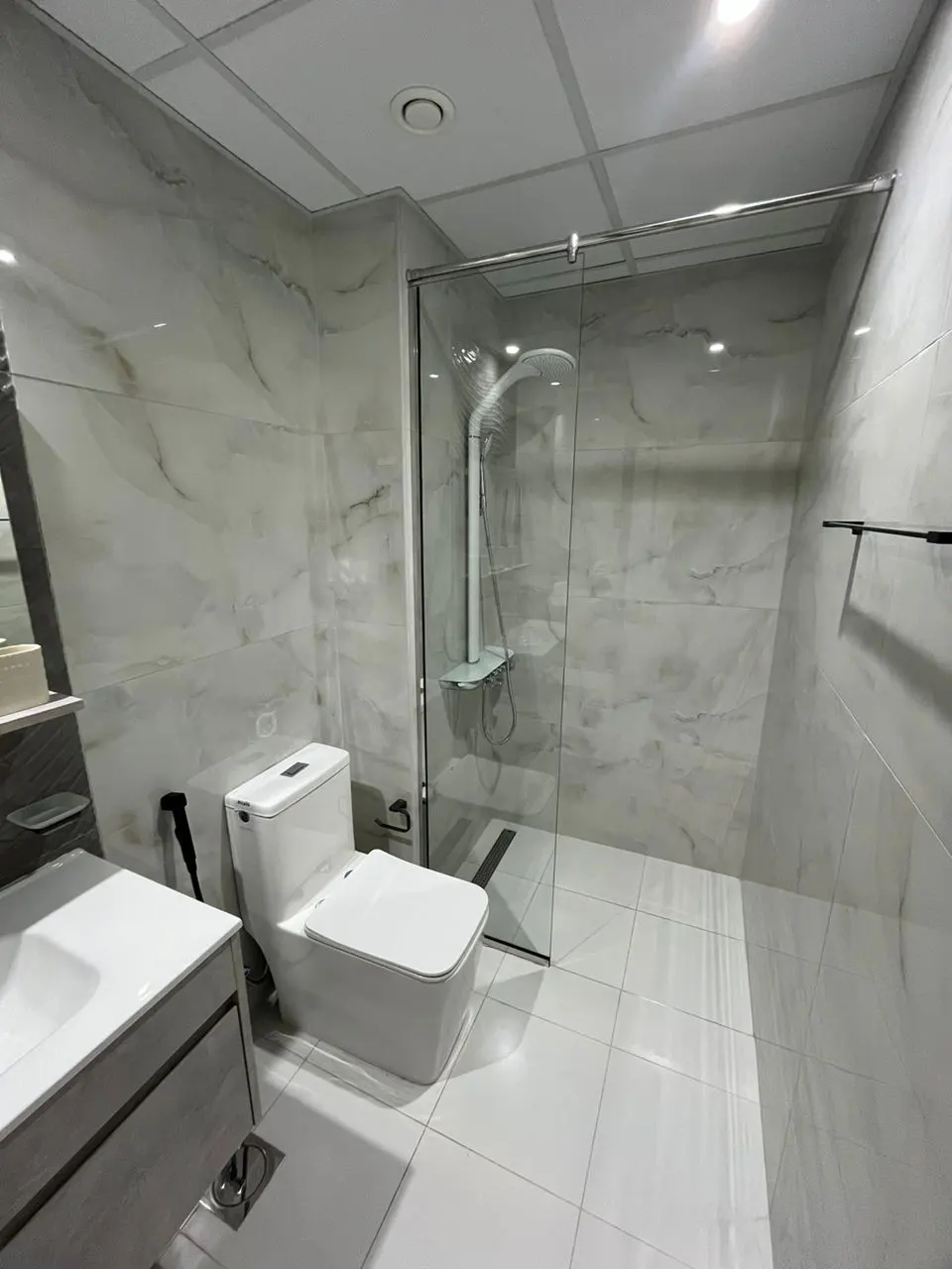 Bright bathroom with white marble walls, white tile floor, glass shower, toilet, and gray vanity.