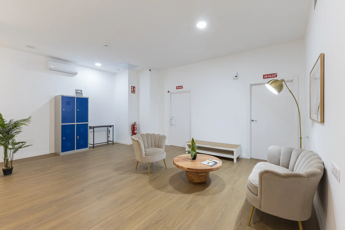 Bright waiting area with two beige chairs, a round wood table, and blue lockers on a wood floor. "En Salida" sign above a door.