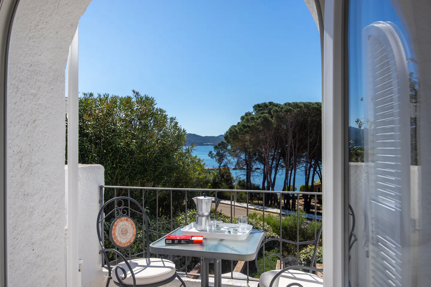 Balcony view of the sea. Table with coffee maker and cups. Chairs with cushions. Blue sky and green trees.
