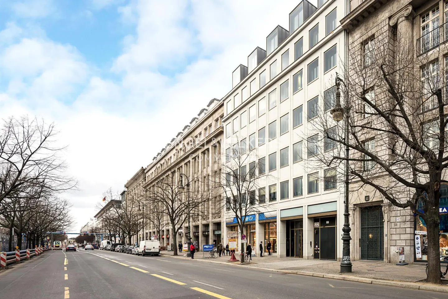 Street view of multi-story buildings with shops, bare trees, and cars on a road under a cloudy sky.