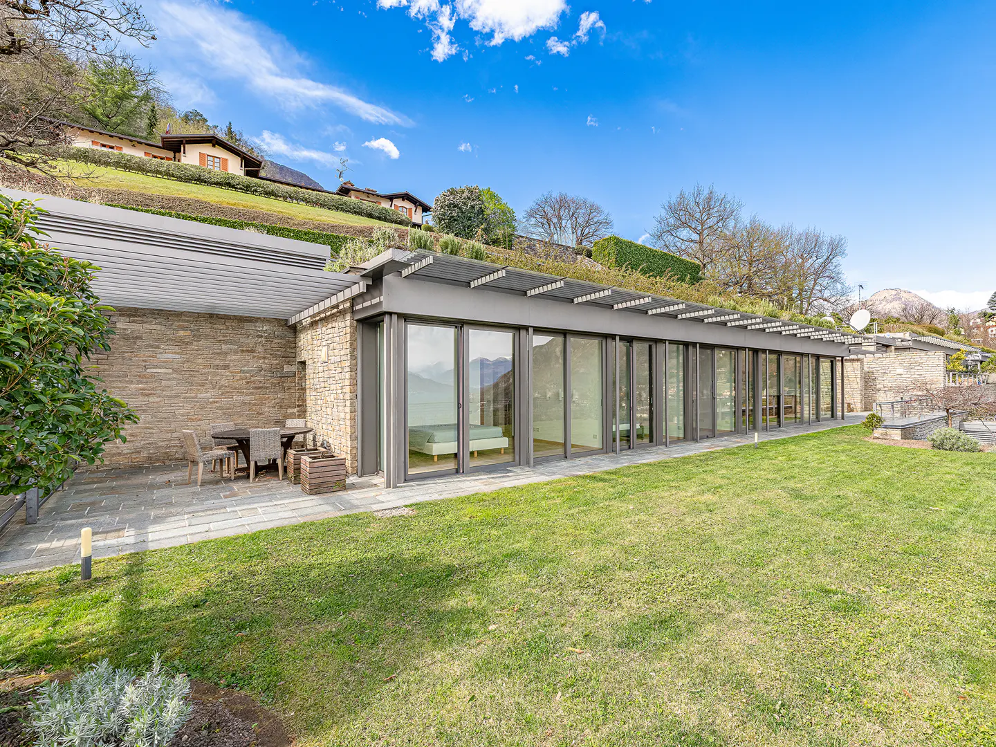 Modern home exterior with stone walls, large windows, and a green roof, set against a grassy lawn and blue sky.