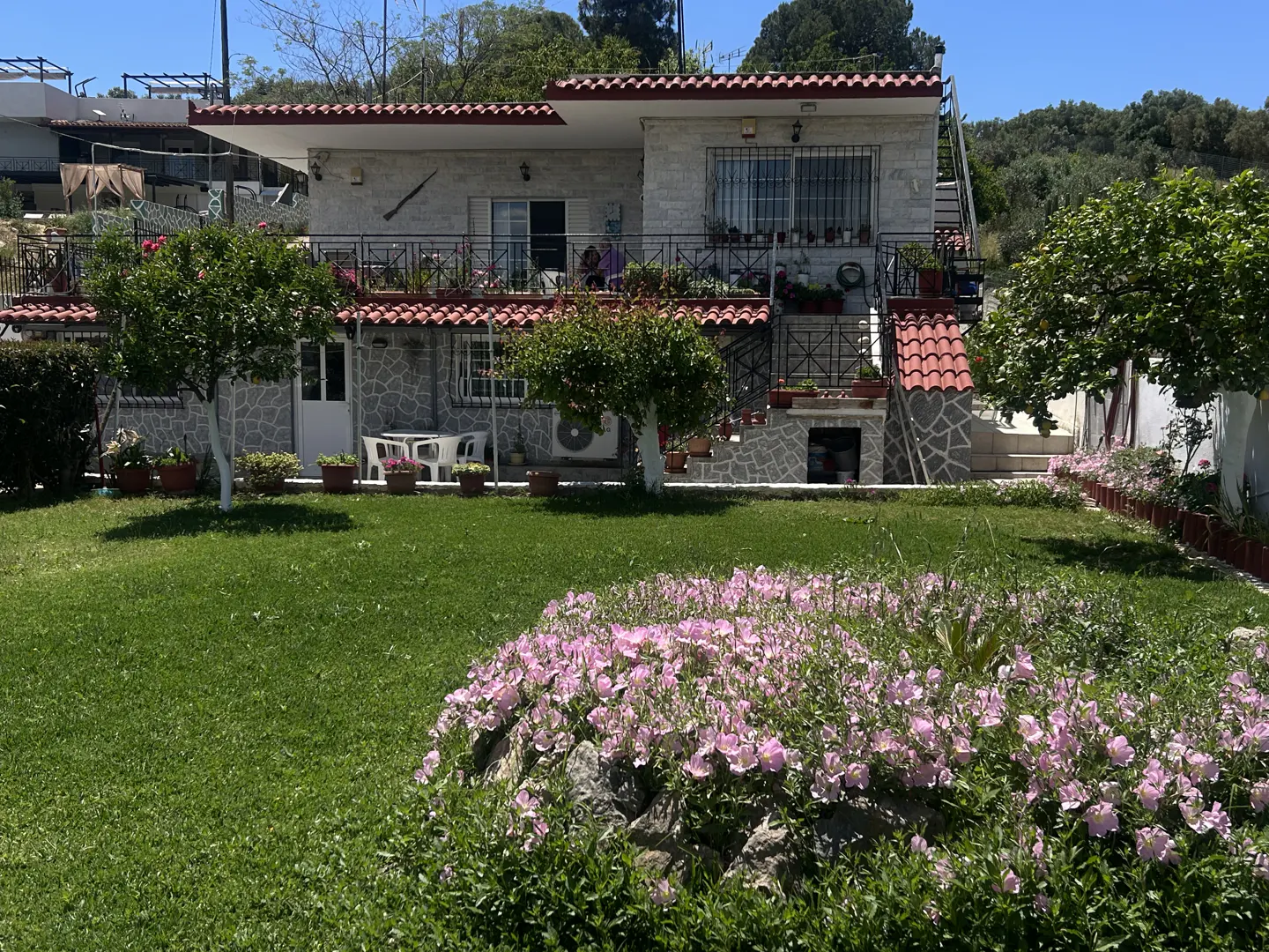 Two-story stone house with a red tile roof, balcony, and lush green lawn featuring a bed of pink flowers in the foreground.