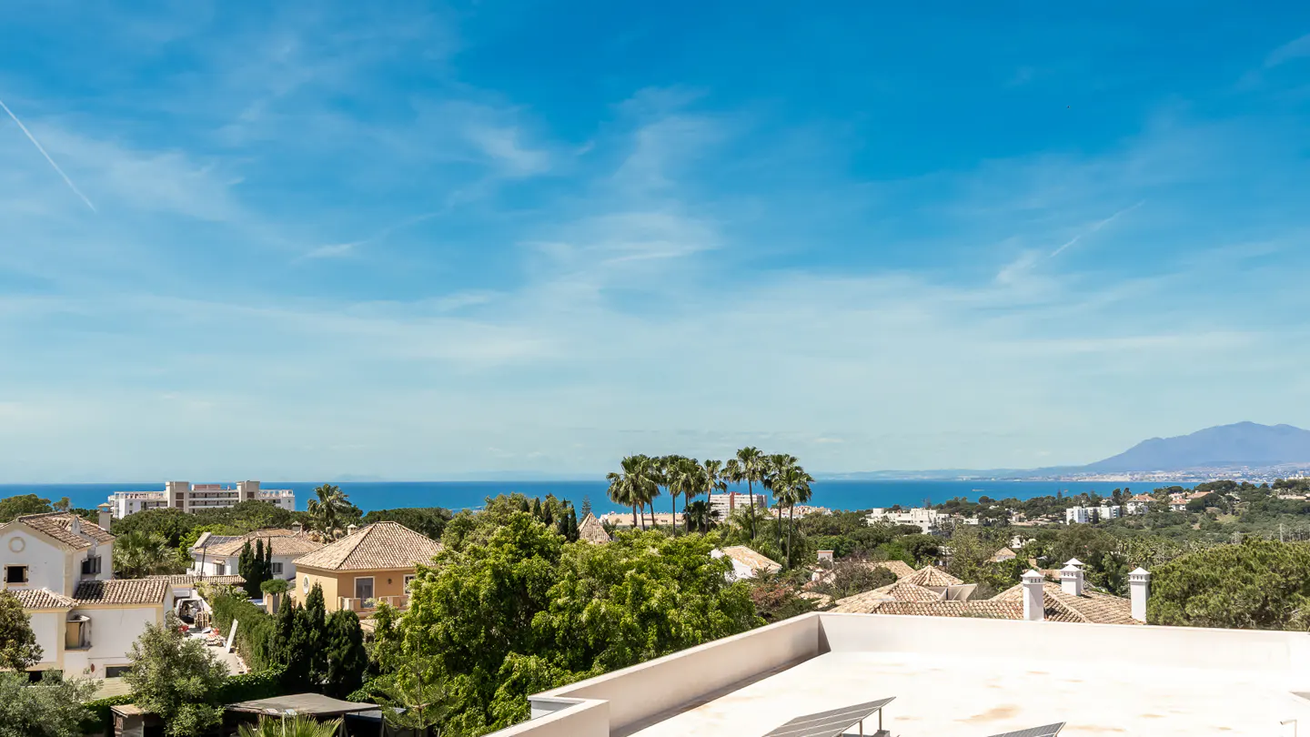 View from a rooftop overlooking houses, trees, and the ocean under a blue sky.