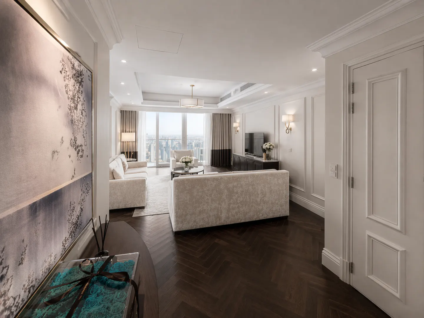 A bright, modern living room with white sofas, dark wood floors, and a city view. A large painting hangs on the wall.