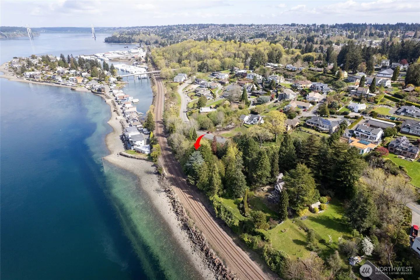 Aerial view of waterfront property with trees, railroad tracks, and a bridge in the background. Blue water and green foliage.