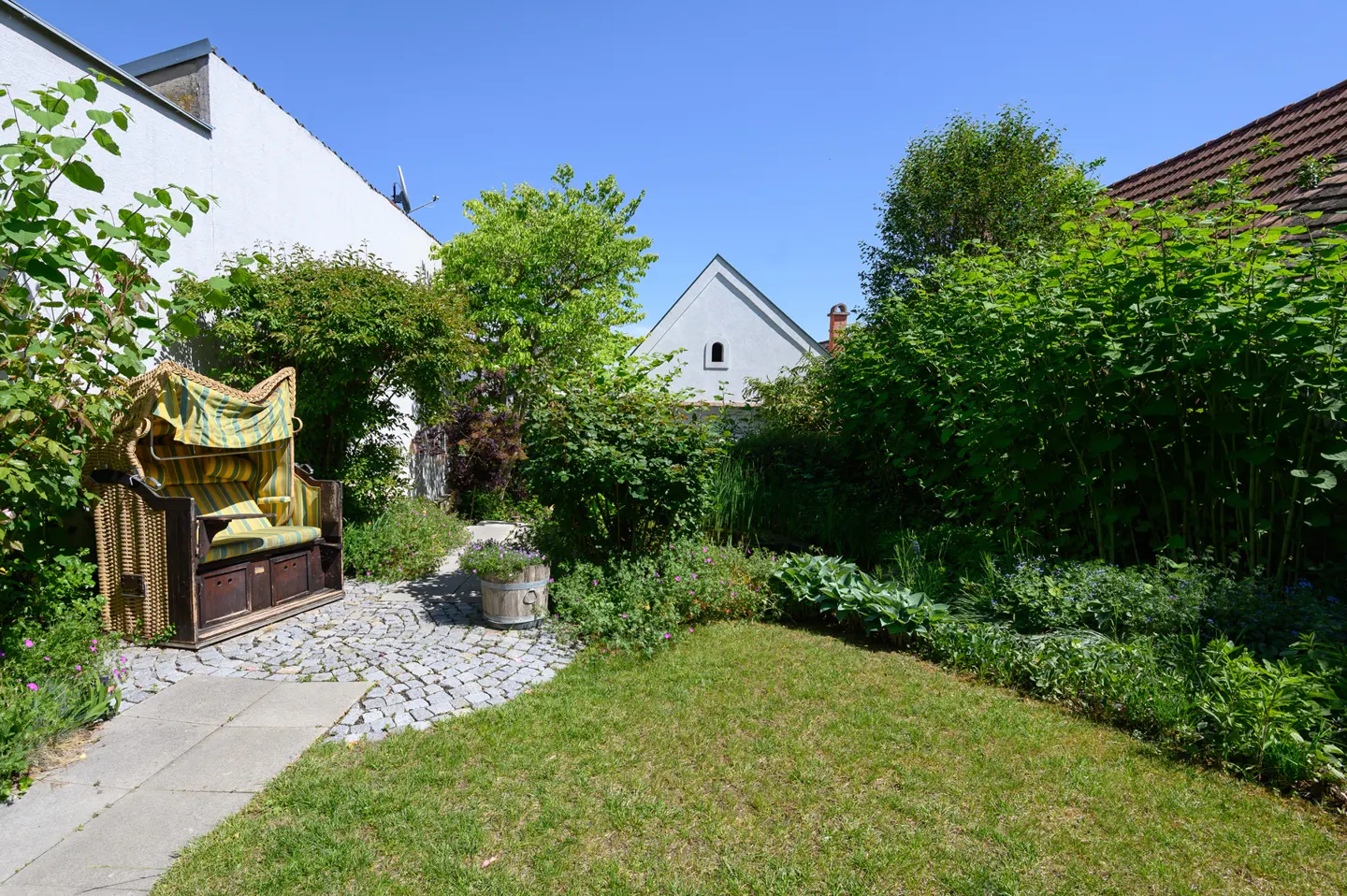 A lush garden with a wicker beach chair on a stone path, surrounded by green plants and grass. A white house is visible in the background.