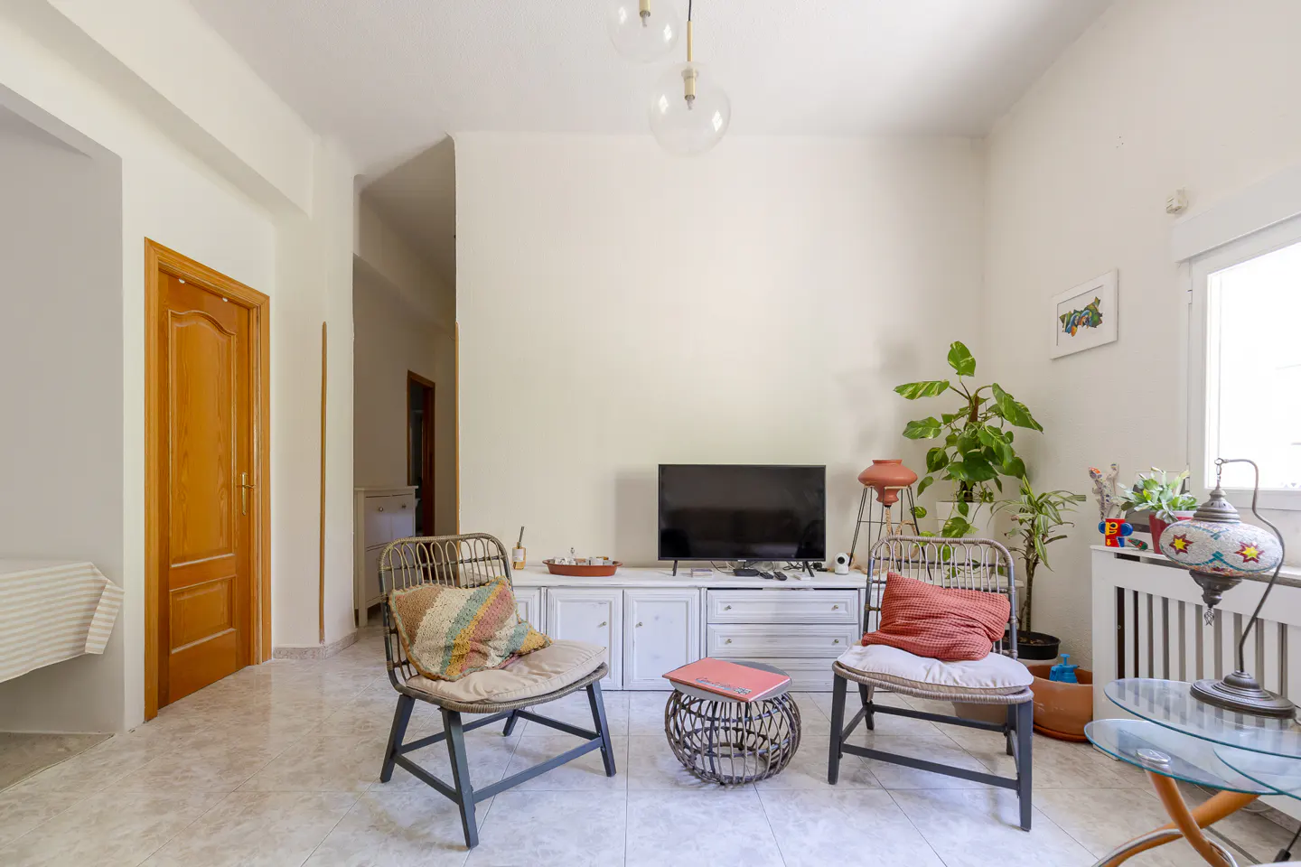 Living room with two wicker chairs, a TV on a white cabinet, and plants near a window. The walls are off-white.