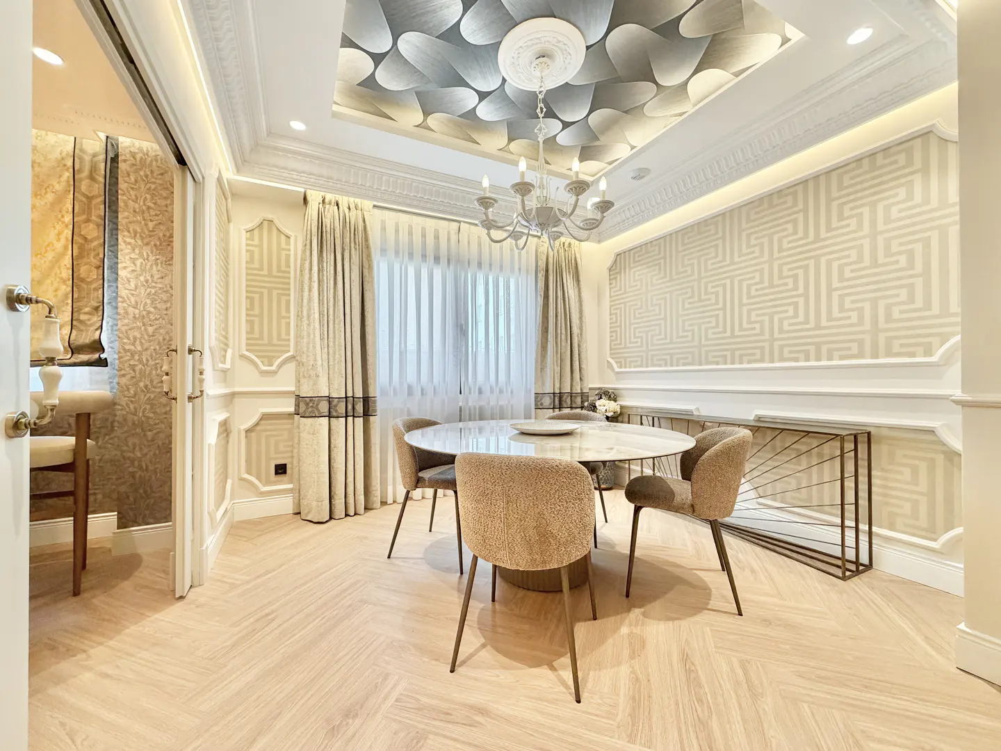A bright dining room with a round table, beige chairs, and herringbone wood floors. The ceiling has a decorative pattern and a chandelier. Curtains frame a window.