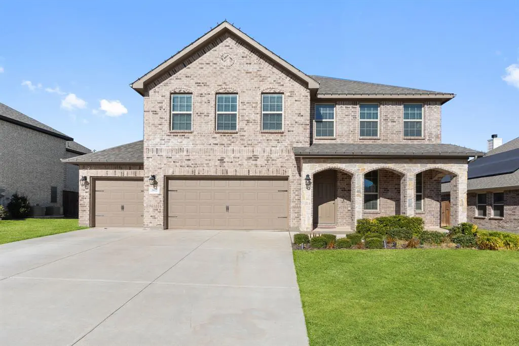 Two-story brick house with a two-car garage, green lawn, and blue sky.