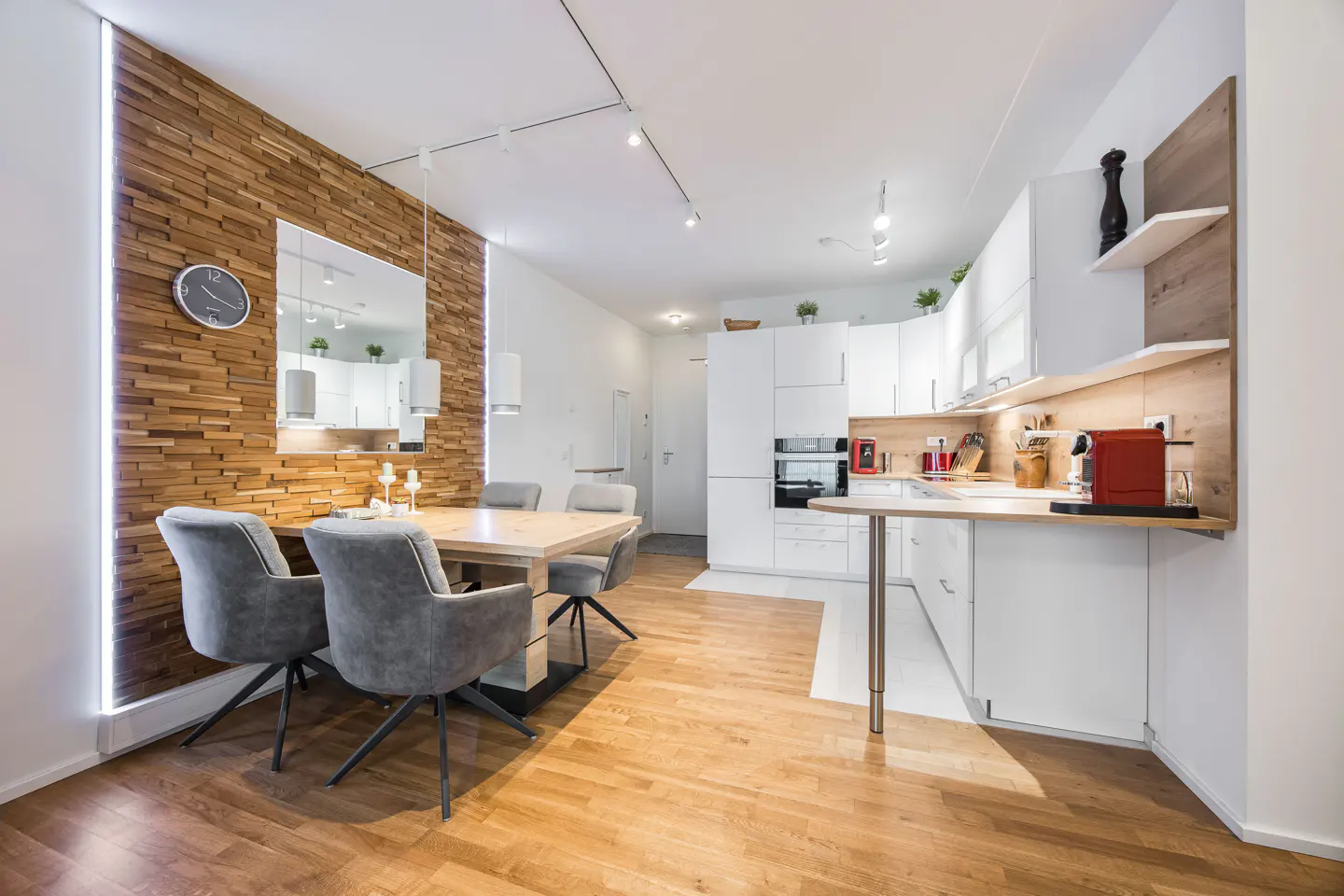 Bright, modern kitchen and dining area with wood floors. A light wood table is surrounded by gray chairs. White cabinets and wood accent wall.