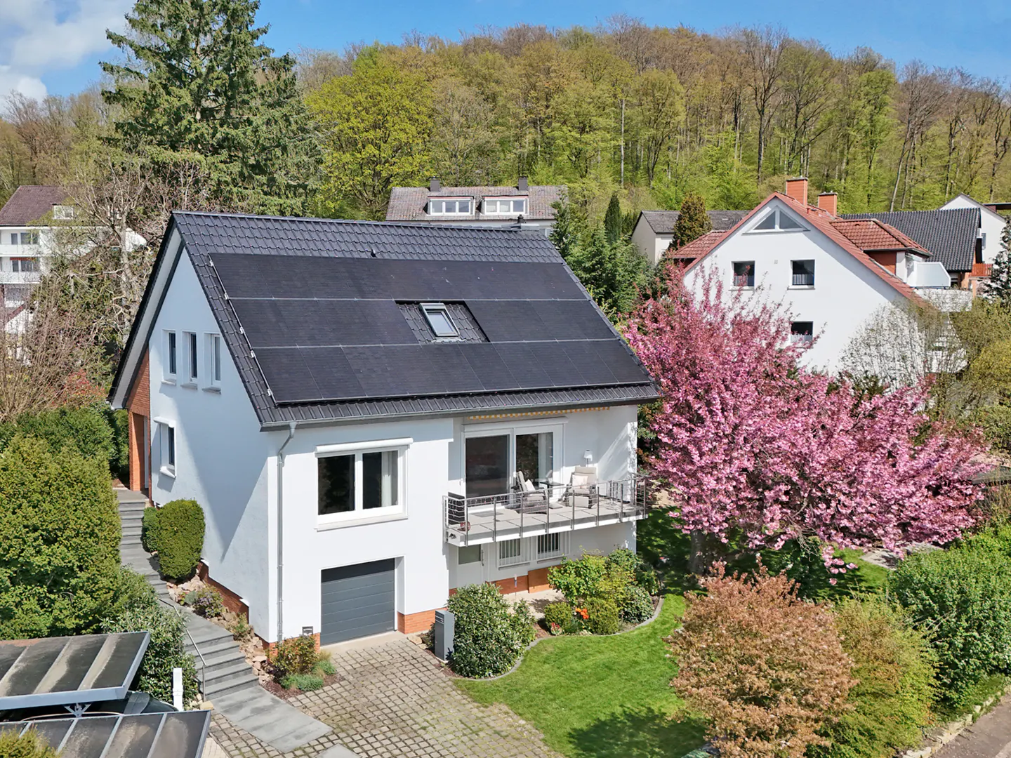 A white two-story house with a dark gray roof and solar panels, surrounded by green trees and a pink flowering tree.