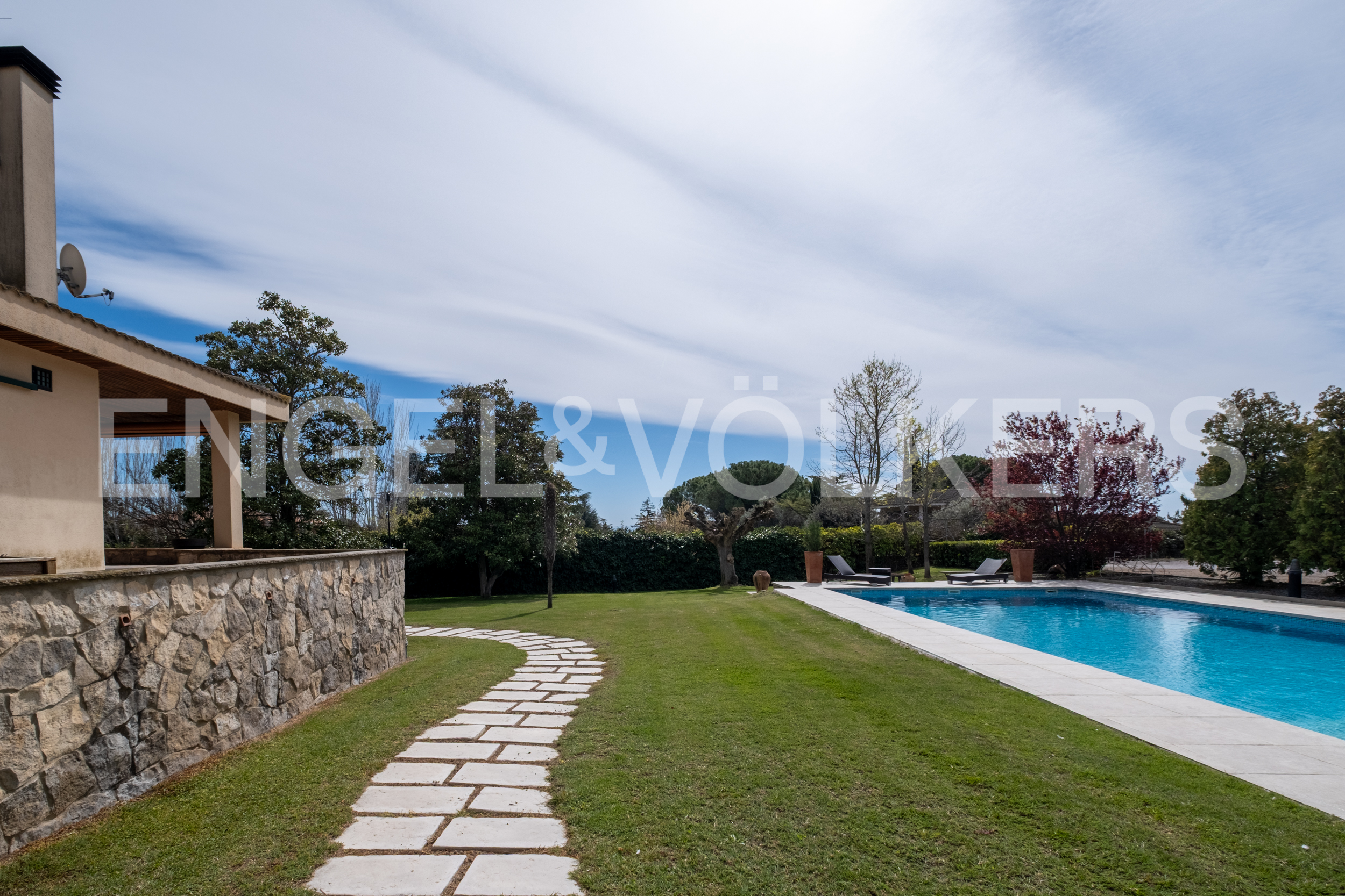 A backyard with a stone path leading to a blue swimming pool, green grass, and trees under a cloudy sky.