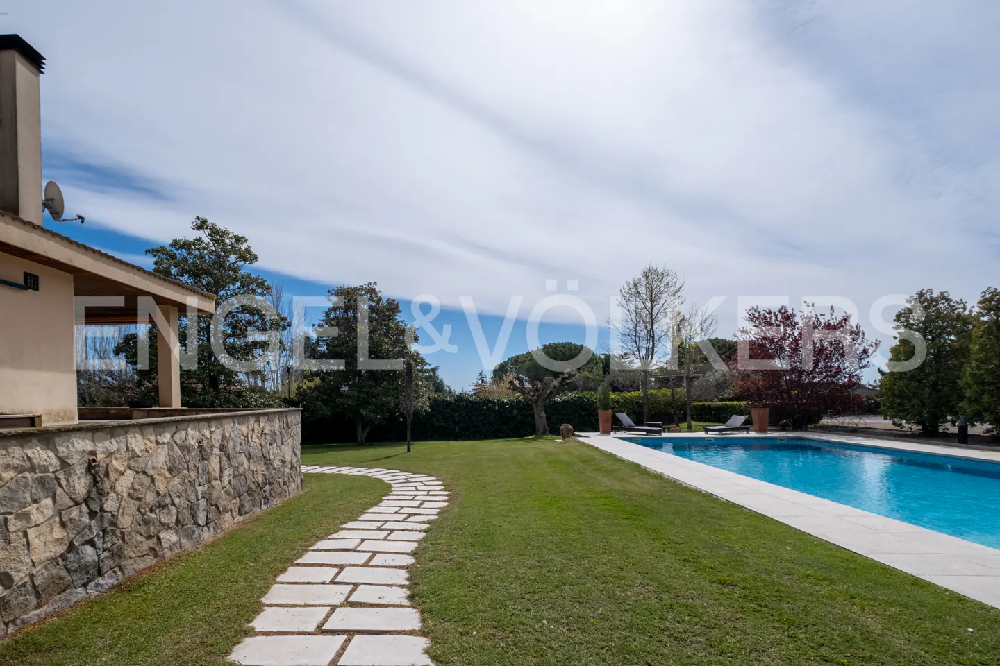 A backyard with a stone path leading to a blue swimming pool, green grass, and trees under a cloudy sky.