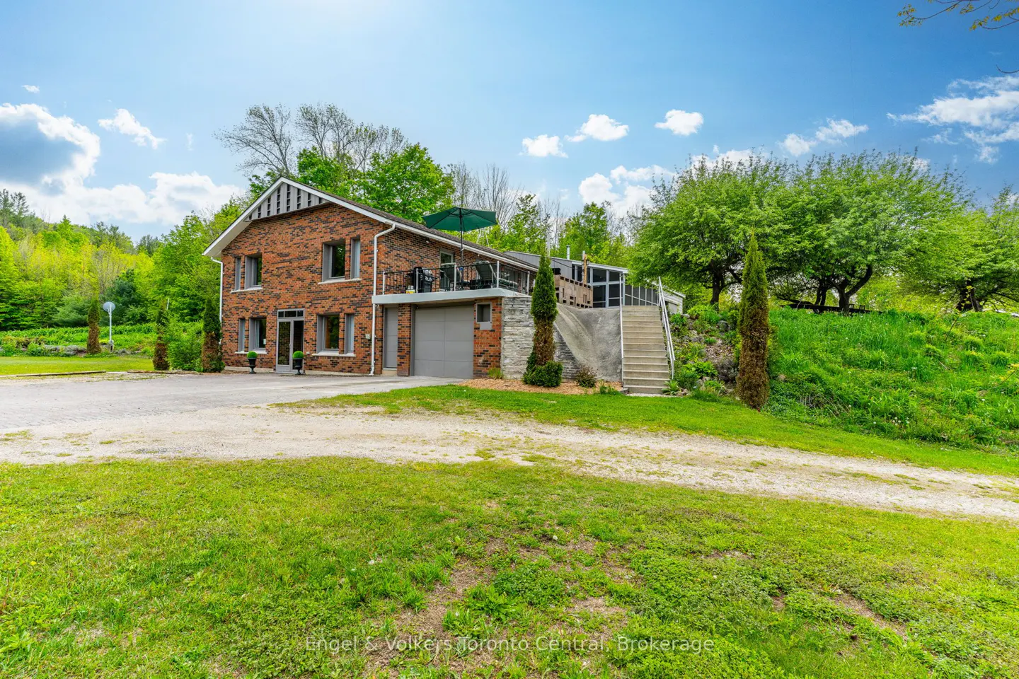 Two-story brick house with a gray garage, a balcony, and a concrete staircase on a grassy hill.