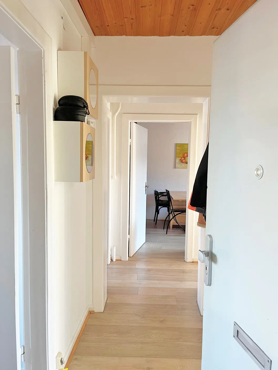 Hallway view with a white front door ajar, light wood floors, and a white wall with two square shelves. A dining room is visible through the doorway.