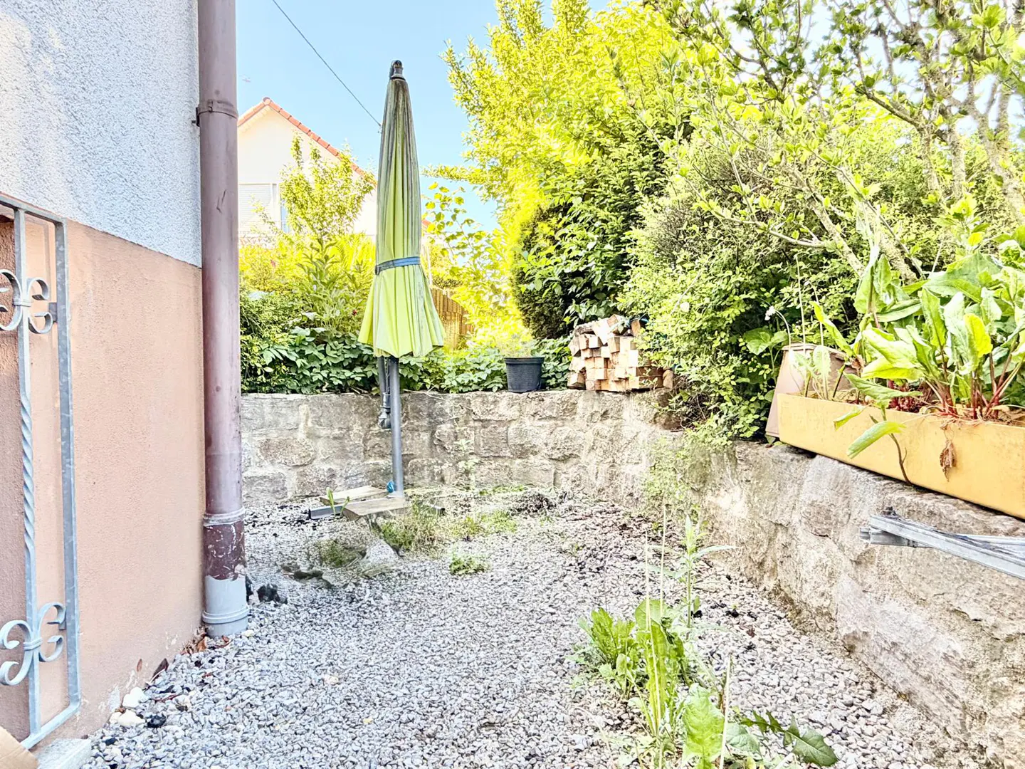A small gravel backyard with a closed green umbrella, stone wall, and greenery.