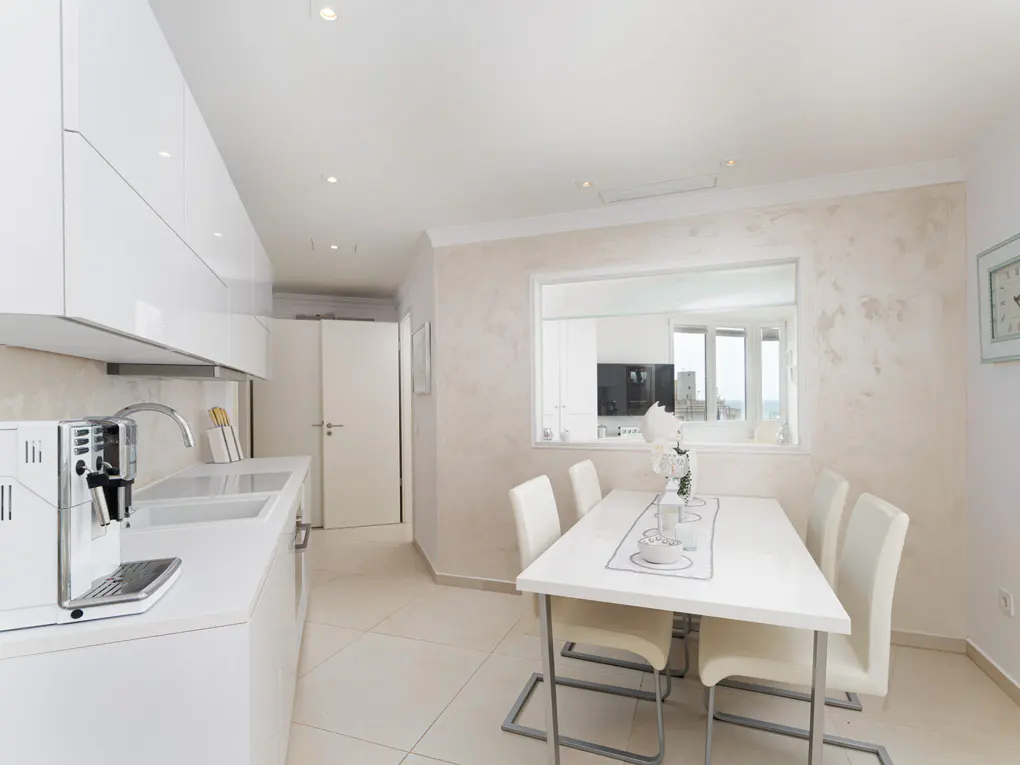 Bright, modern kitchen with white cabinets, countertops, and dining table with six chairs. A coffee machine sits on the counter.